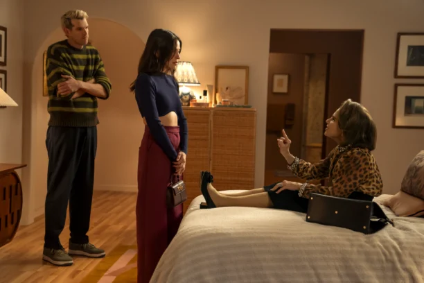 NWT 303 Unit 01491RC2.jpg "Three people in a cozy, warmly lit bedroom; one woman sits on the bed gesturing, another stands with a purse, and a man in casual clothes stands nearby, watching the conversation."