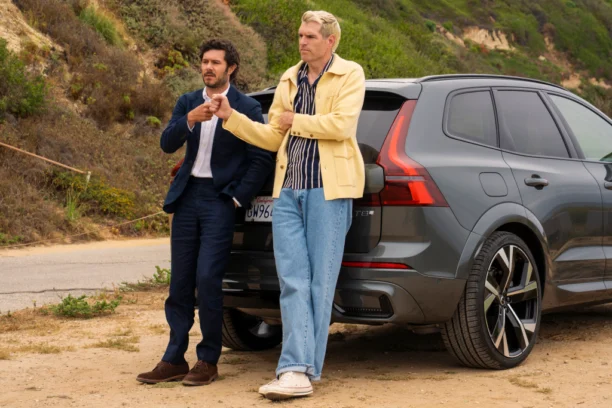 NWT 310 Unit 01641RCX.jpg "Two men in casual and formal clothes stand outside, leaning against the back of a gray SUV parked on a dirt road, with green hills and vegetation in the background, appearing to have a conversation."