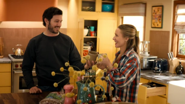 Nobody Wants This n S3 E1 00 04 05 08RC2.jpg "A man and woman smile and toast with mugs in a cozy, sunlit kitchen, surrounded by yellow cabinets, kitchen appliances, and decorative items, creating a warm, homey atmosphere."