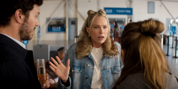 Nobody Wants This n S3 E4 00 12 56 21RC.jpg "Three people talking in an airport terminal, one woman in a jean jacket with fur collar gesturing animatedly, appearing to explain something to a man holding a drink and a woman with her back to the camera. Signs and check-in lines in background."
