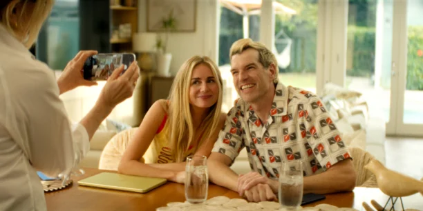 Nobody Wants This n S3 E6 00 03 12 00RC2.jpg "A smiling couple sits at a table indoors, holding hands and posing for a photo taken by another person. Bright, cozy setting with large windows, plants outside, two glasses of water, and a yellow tablet on the table."